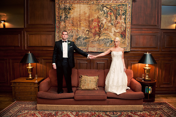Bride in champagne a-line dress and groom in black and white tuxedo standing on a dark red and gold sofa while holding hands - photo by Houston based wedding photographer Adam Nyholt